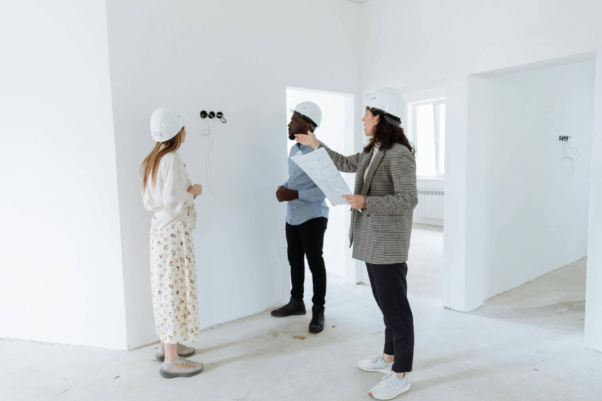 Three individuals stand in an empty room surrounded by various construction equipment, highlighting a work-in-progress environment.