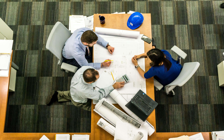 A group of three people engaged in a discussion over blueprints at a table, focused on their project.